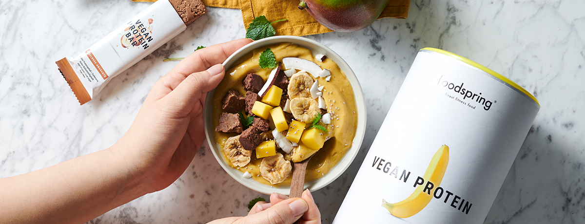 A pair of white-presenting hands holds a white stoneware bowl filled with a vitamin D rich, yellow, vegan protein smoothie bowl topped with banana chips, mango cubes, coconut chips, and vegan protein bar chunks