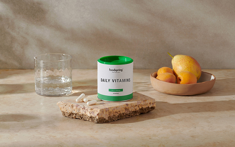 A canister of Daily Vitamins by foodspring stands on a slab of stone.