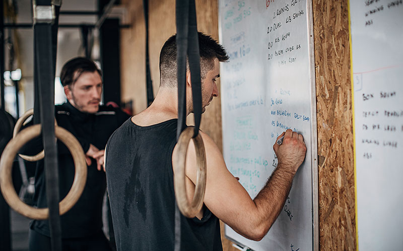 A white man writes a workout plan on a whiteboard as another white man looks on.