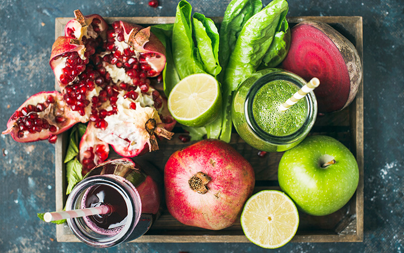 A tray of smoothies scattered with pomegranates, spinach, a lime, a red beet, and a green apple