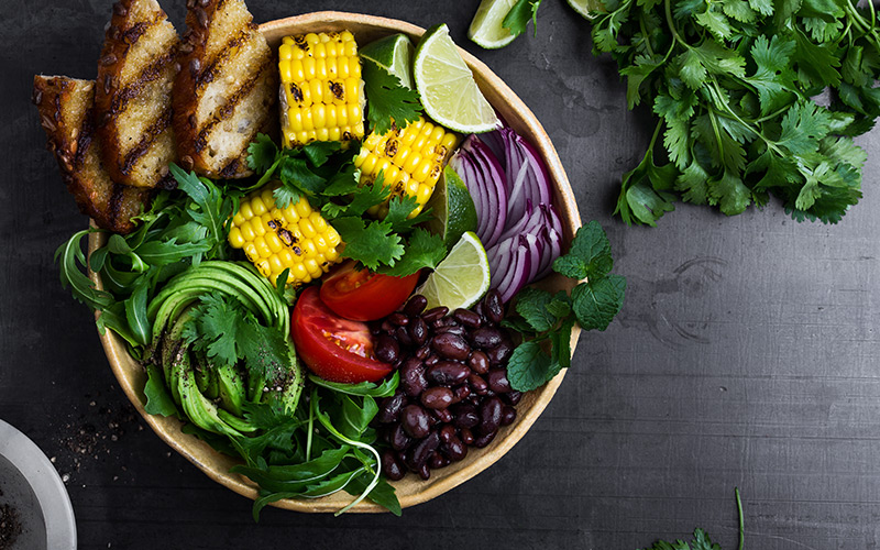 A bowl of deeply colored vegetables, tomatoes, corn, chicken strips with black grill marks, kidney beans, red onions, and green salad leaves sits on a gray background