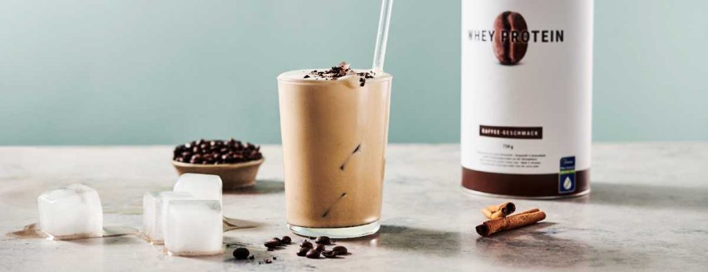 A glass of iced coffee protein shake sits with a glass straw sticking out of it. In front of the glass are a few coffee beans. Behind the glass, a canister of Coffee Whey Protein.