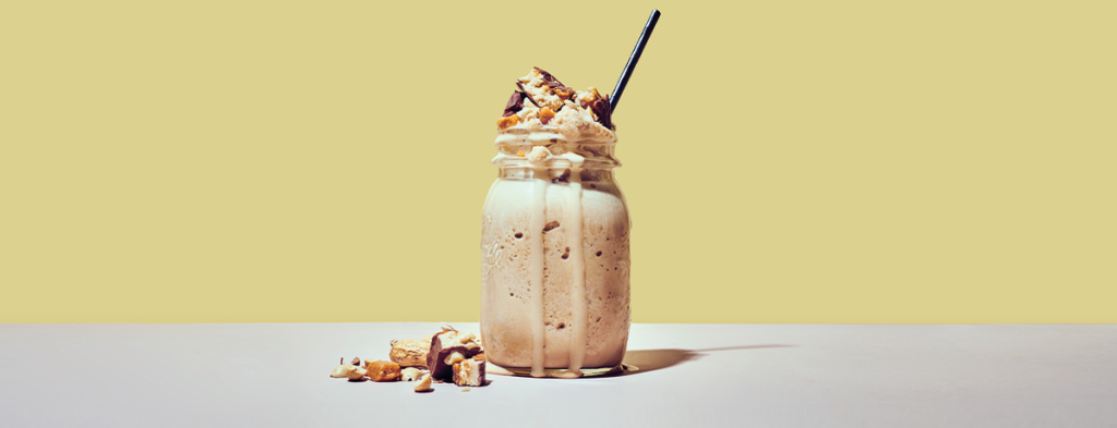 A photo of a coffee peanut protein shake in a Mason jar, topped extravagantly with cubed chocolate peanut protein bar and chopped peanuts