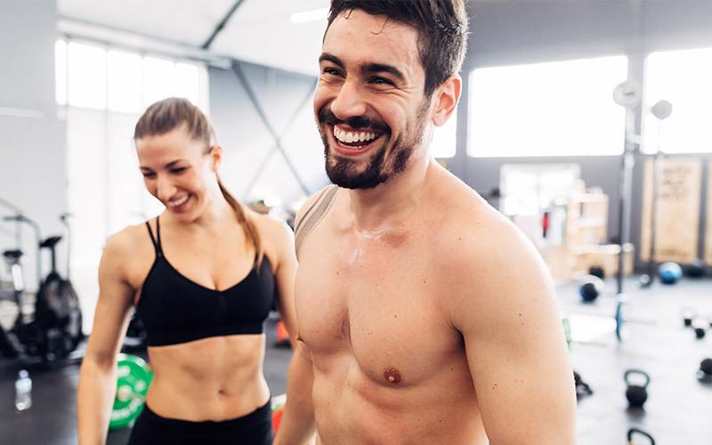 A smiling white man and woman prepare to do a couple workout in an indoor gym setting