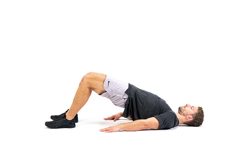 A short-haired white man performs a glute bridge in front of a white background.