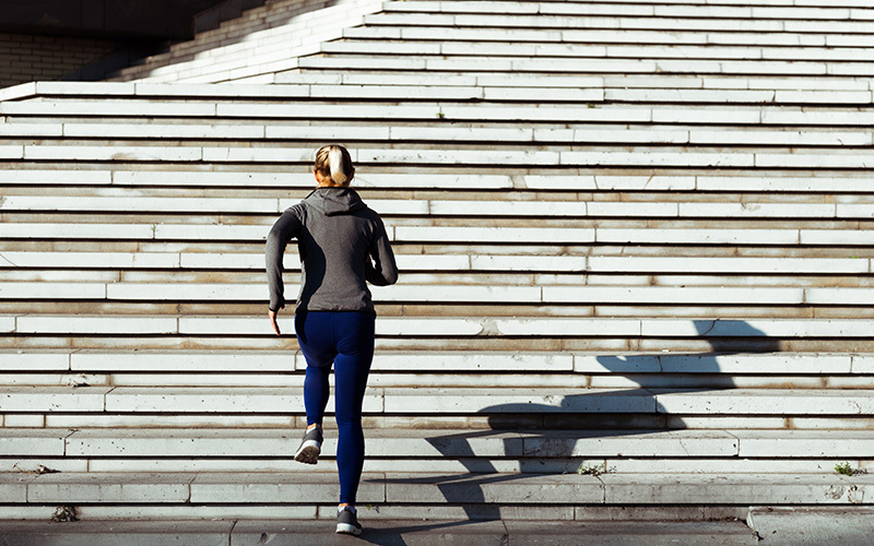 How to start jogging - a woman seen from behind with a long blonde ponytail, a gray hoodie and blue leggings. She appears to be jogging up from the bottom of a set of white concrete stairs outdoors.