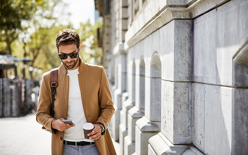 A man with short brown hair walks past a gray-walled building while looking at a phone and holding coffee in a to-go cup