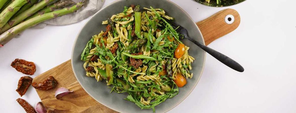A green and fresh-looking bowl of pasta salad with asparagus, garnished with yellow cocktail tomatoes, dark red sun-dried tomatoes, and fresh green arugula leaves