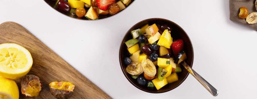 A black bowl holds bright, refreshing fruit salad with a spoon stuck into ot