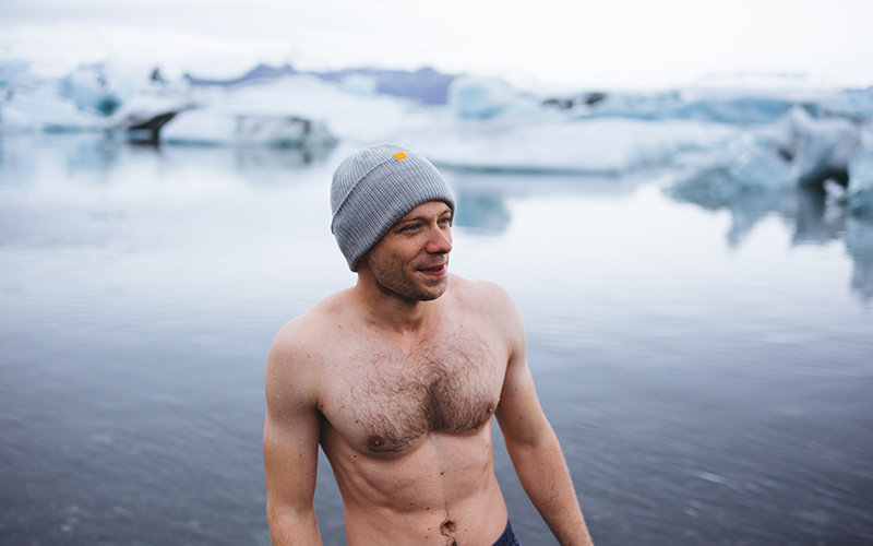 Mann steht Oberkörperfrei und mit Wollmütze vor einem fast zugefrorenen See