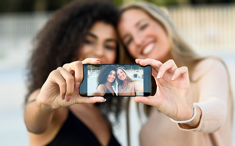 Two women of different races take a selfie on a phone. The viewer is able to see the people's faces on the screen of the phone in focus, rather than the faces of the actual people.
