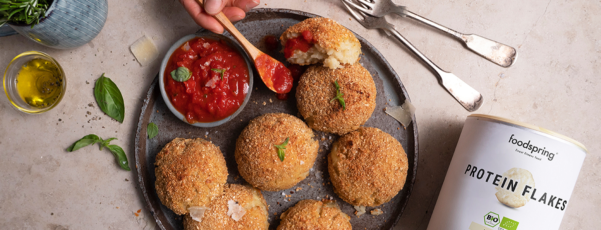 a slate-gray plate of fitness arancini sits with a small bowl of deep red tomato sauce on the plate. A few medium-skin-toned fingers invitingly hold a wooden spoon scooping some of the sauce out.