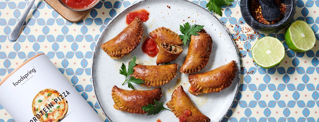 A white plate of medium-browned protein empanadas garnished with greens and dollops of red tomato salsa