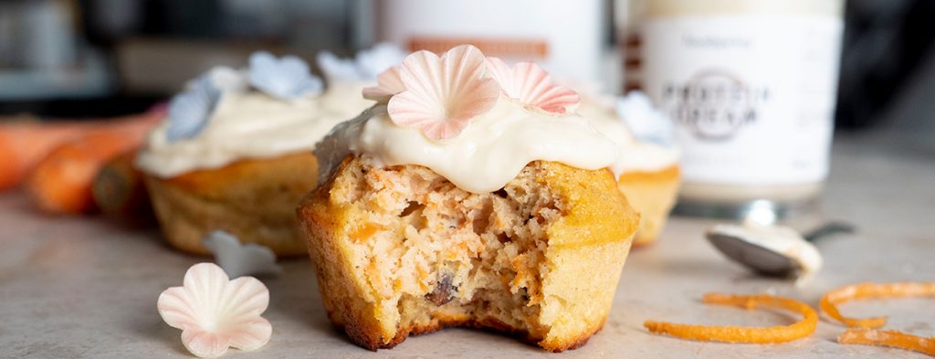 A carrot cake muffin topped with a dollop of white frosting, with a bite taken out of it to show the moist interior