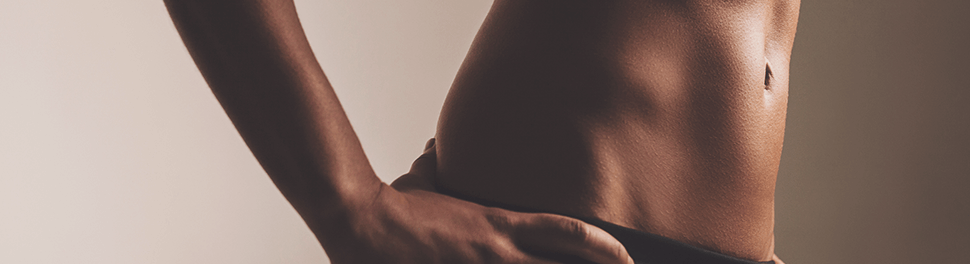 A profile view of a medium-skin-toned, slim stomach area, probably of a woman, with smooth skin. Part of the model's hand is visible resting on their front hip.