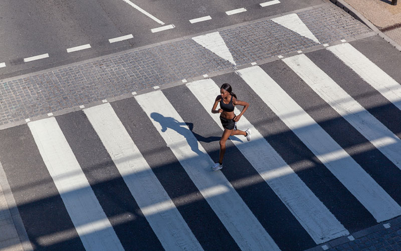 a woman of color runs for endurance training as she crosses a road on a large zebra-striped crosswalk. She is seen from a distance, from above.