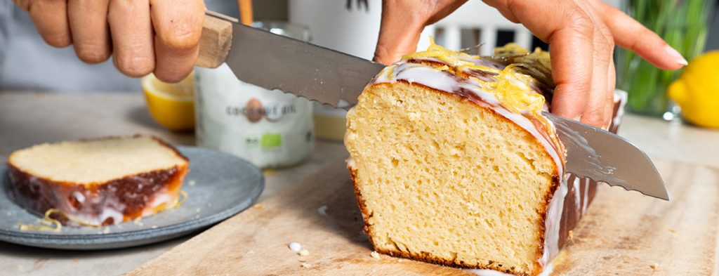 a medium-skin-toned hand holds a lemon cake gently while using a bread knife to slice off a piece.