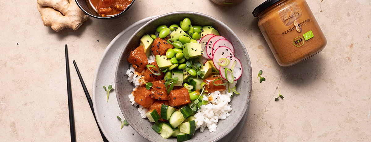 A photo from above of a Poke Bowl filled with rice, edamame, avocado cubes, marinated raw fish, radish slices, and cucumber chunks with a jar of foodspring Peanut Butter next to it.