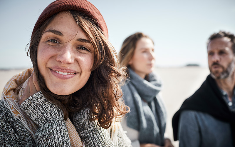 A white woman smiles at the camera. She wears a hat. The two people and the background behind all of them are out of focus, but it's clear they are outdoors as there's a blue sky behind their heads.