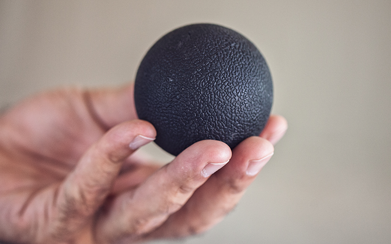 A white-skinned hand holds a black fascia ball with a gentle snakeskin texture visible