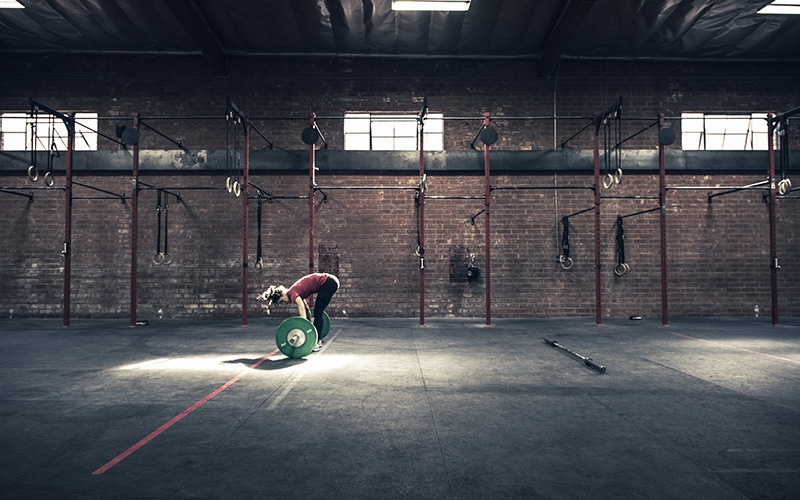 A white woman stands in an empty, large gym setting, bending at the waist to pick up a barbell holding heavy weights