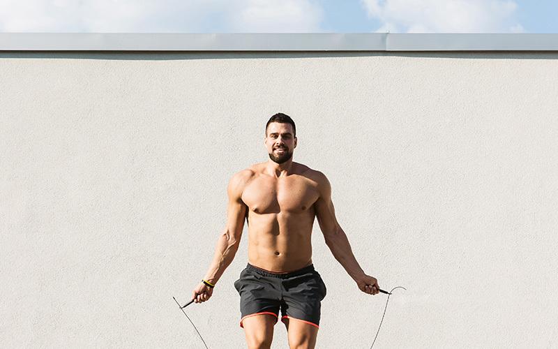 a shirtless, muscular man performs a jump rope workout outdoors in front of a cream-colored wall