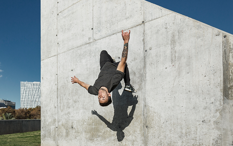 A white man performs a parkour somersault, running up a wall and flipping over backwards. The photo has captured him fully upside-down with one foot still against the wall. 