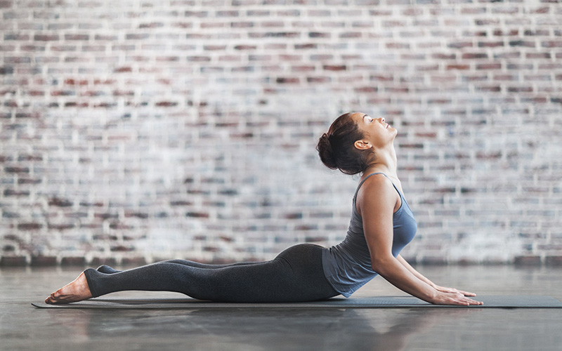 A white woman does a cobra yoga position on a mat in front of a brick wall indoors.