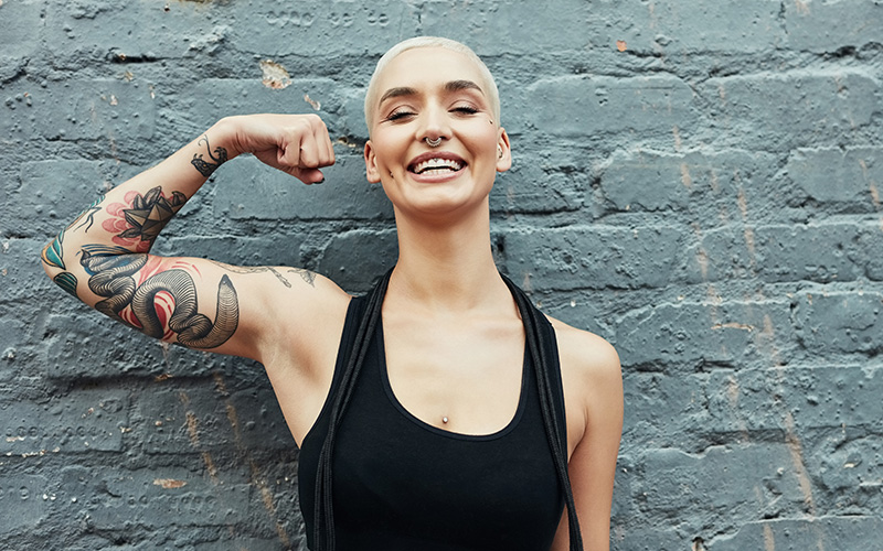 A white woman with a buzz cut flexes her tattooed right arm showing a smile of resilience