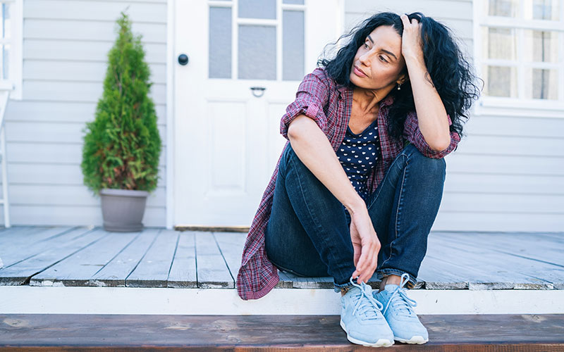 An older, dark-haired woman who may be experiencing or may be thinking about menopause. She sits outdoors on the porch of a white-washed house.