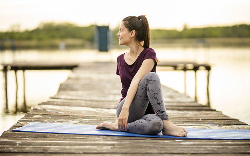 A white woman stretches her back on a blue yoga mat on a pier