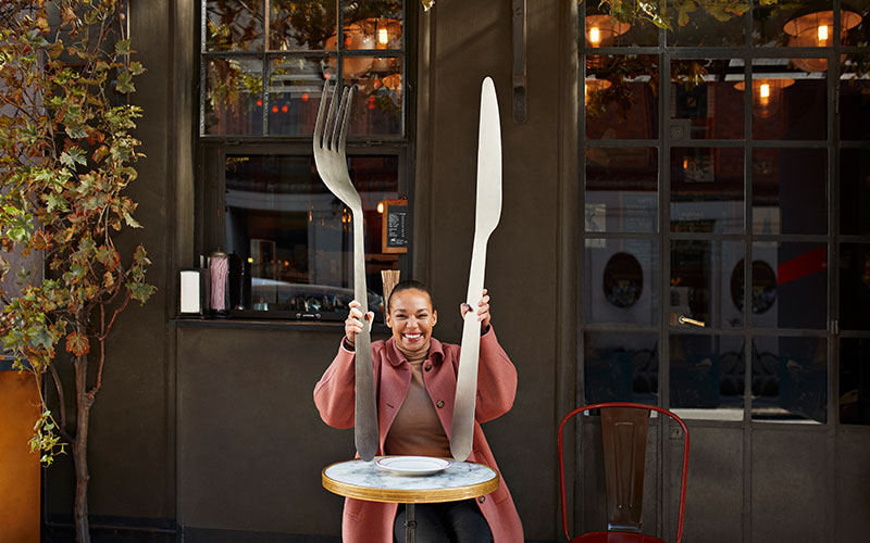 Frau sitzt mit Lebensgroßen Messer und Gabel an einem Tisch