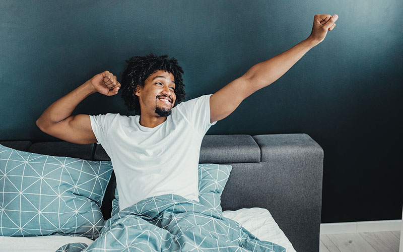 A man of color stretches his muscular arms in bed, displaying the connection between sleep, weight loss, and muscle gain