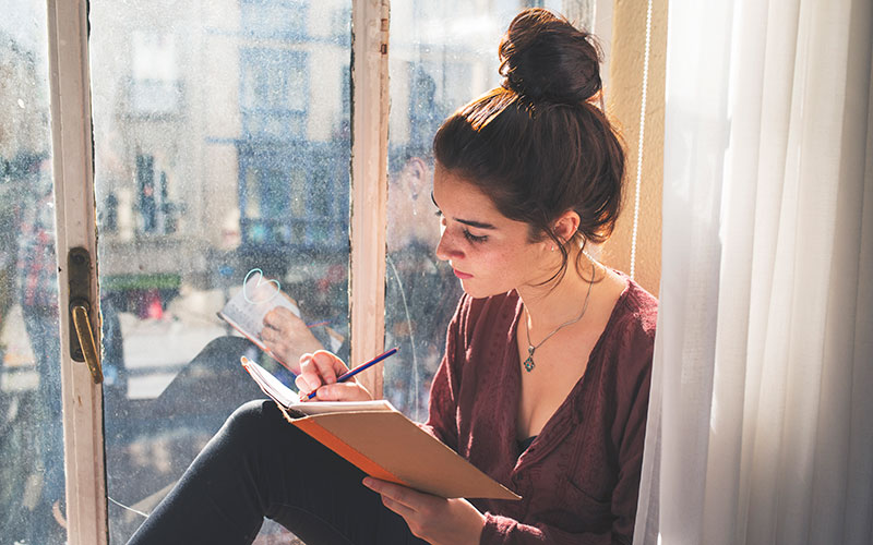Eine Frau sitzt am Fenster und schreibt in ihrem Journal