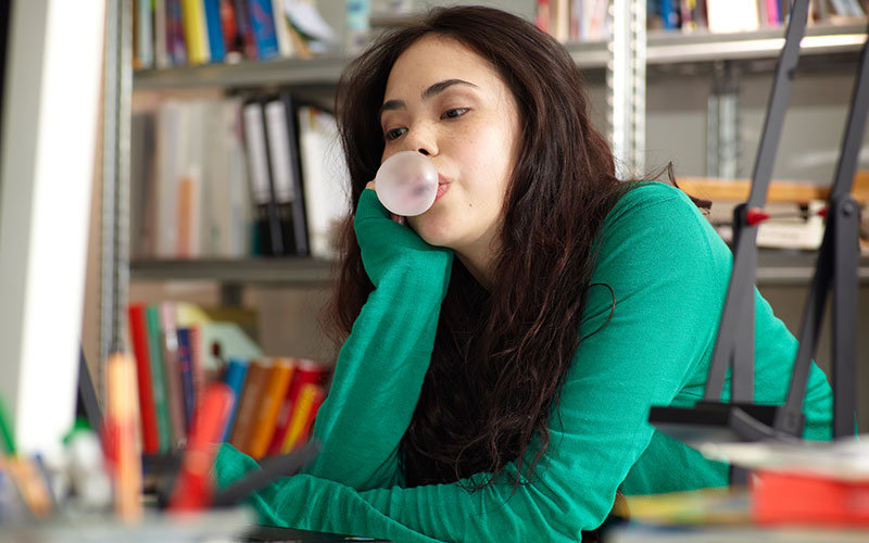 A white, dark-haired woman blows a bubble while sitting at a desk, hoping to stop her procrastination