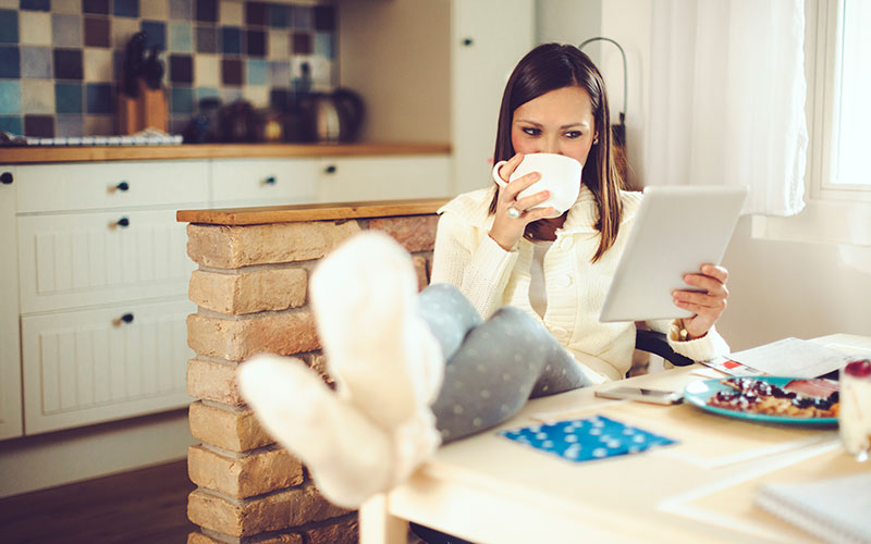 A brown-haired white woman rests her slippered feet on her kitchen table, sipping from a mug of tea and checking out a tablet for self-care