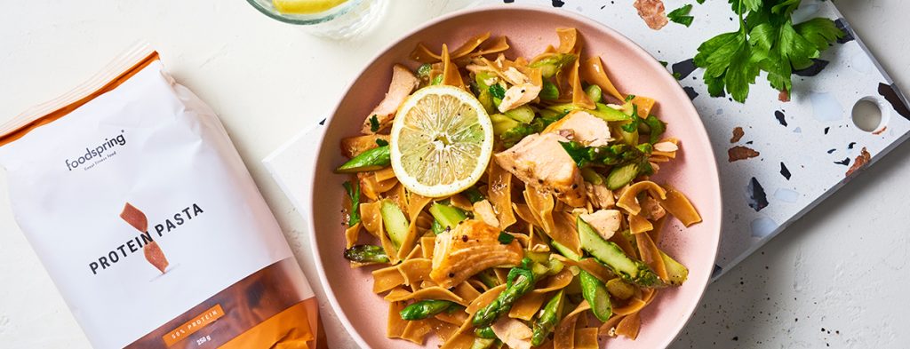 a salmon-pink-colored bowl filled with pasta with salmon and asparagus, garnished with a moon of fresh lime