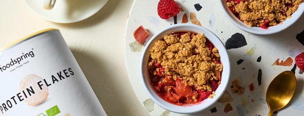 A white ramekin holds a pink-red portion of low-sugar rhubarb crumble topped with golden-brown streusel