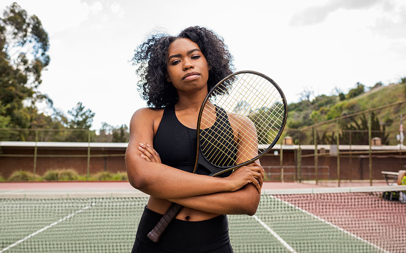 Frau steht mit Tennisschlägern in den Armen auf einem Tennisplatz