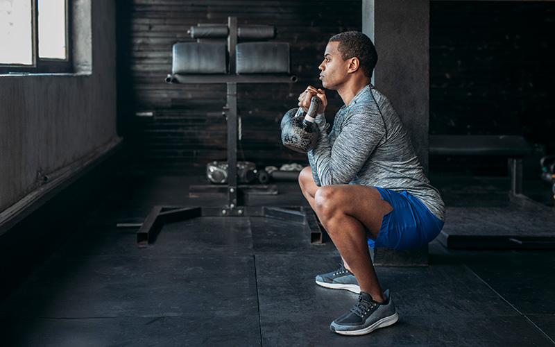 A man of color in sports gear does a goblet squat indoors, holding a kettlebell.