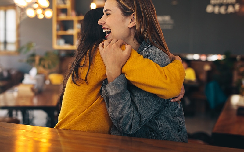 Two long-haired feminine-presenting people hug. One is white, and the other's skin is fully out of the camera angle. The white woman is smiling with joy and gratitude.
