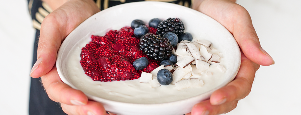 two white hands hold a bowl of yogurt topped with fresh berries, coconut chips, and chia cherry jam