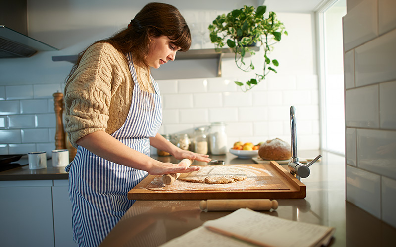 A woman rolls out dough using one of these top baking hacks. she works on a wooden cutting board, with a white-tiled home kitchen wall behind her.