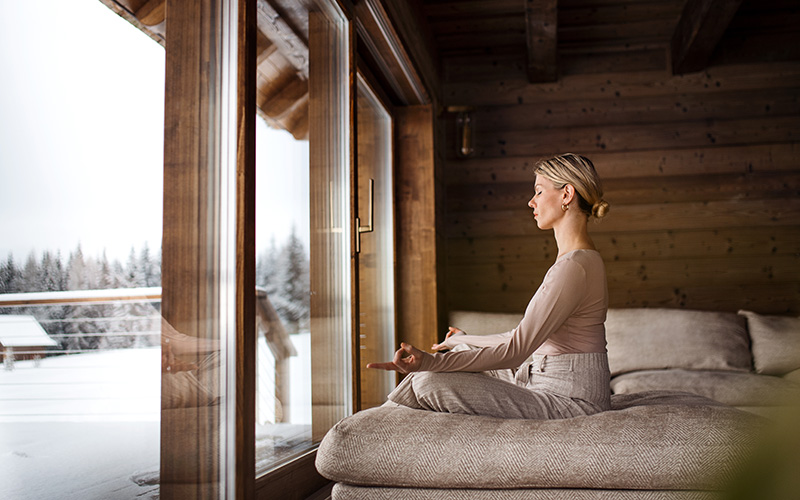 A white woman sits on a bed, meditating towards her window after a winter yoga session, looking out on the winter landscape outside her window.