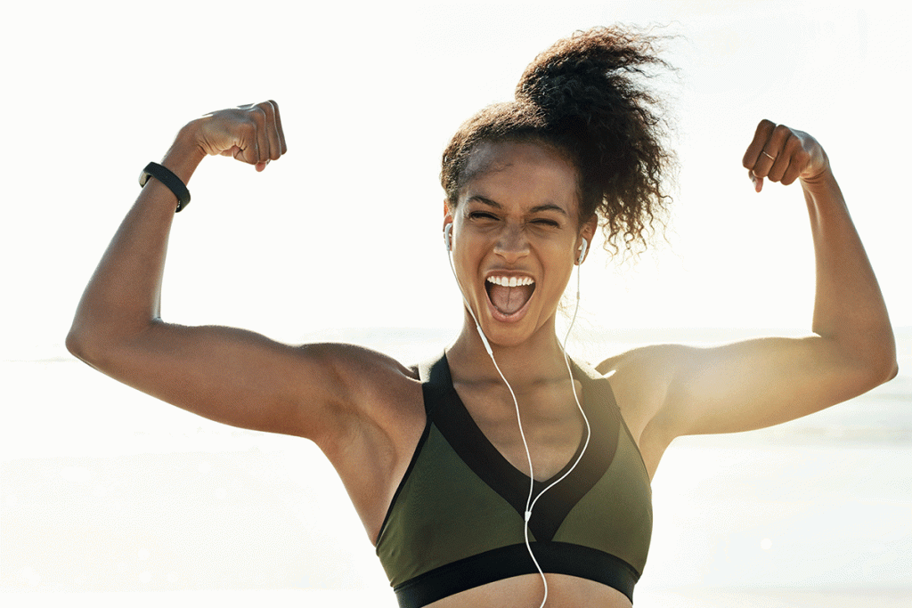 A woman of color flexes her biceps while smiling at the camera, showing how what we do and don't do after a workout and how it affects your body