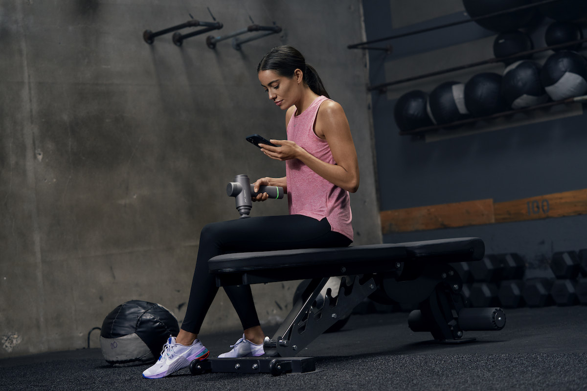 Femme assise sur un banc de musculation à la salle de sport utilisant son téléphone et l'Hypervolt 2