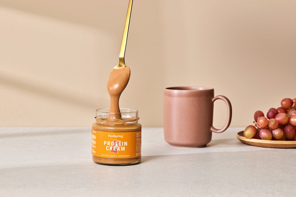 A golden spoon drizzles a line of Salted Caramel Protein Cream into its jar while preparing salted caramel recipes