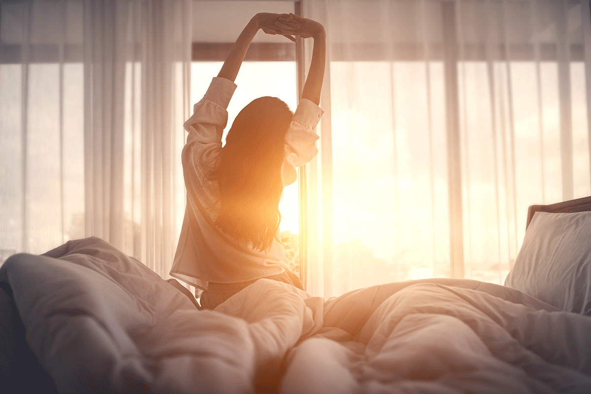 Frau streckt sich nach dem Aufstehen im Sonnenlicht vorm Fenster