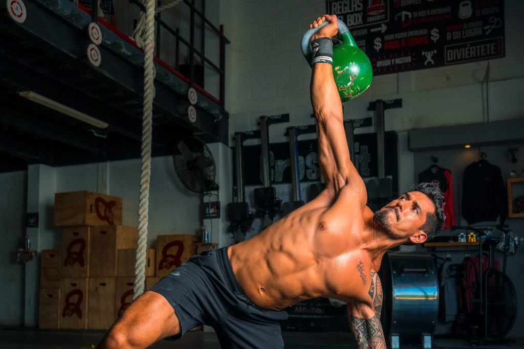 A man is working out with a kettle bell with six-pack abs. It looks like he is in the middle of a Turkish get up.