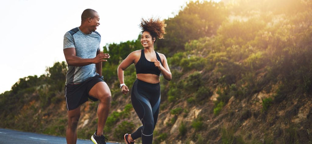 A man and woman of color smile at each other as they run outdoors to improve mental fitness